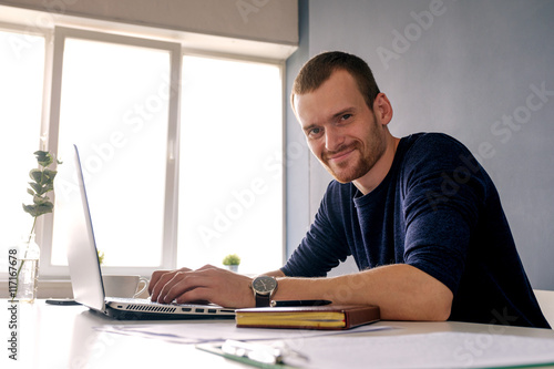 Man in pullover smiling at camera while working on laptop