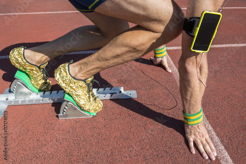 Athlete crouching at the starting line of a running track listening to motivational music on headphones from his mobile phone armband with Brazil colors wristbands 