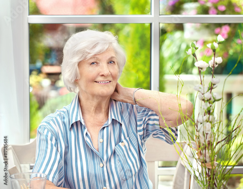  Seniorin sitzt im Wintergarten ,eine Hand im Nacken, im Hintergrund der Garten
