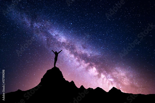 Landscape with vibrant Milky Way. Colorful night sky with stars and silhouette of a standing sporty girl with raised-up arms on the mountain peak on the background of beautiful galaxy.