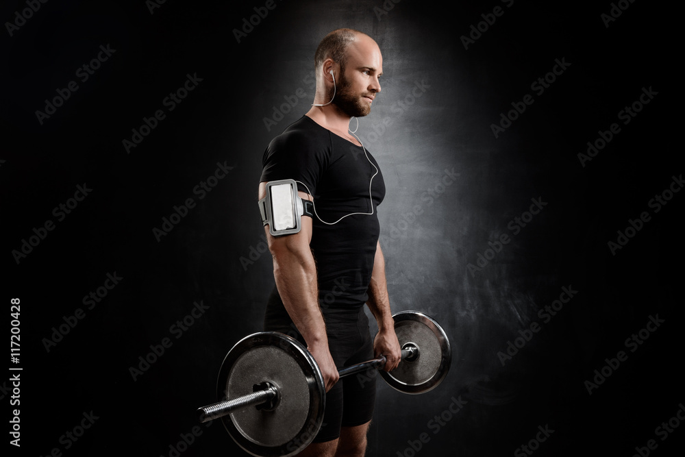 Obraz premium Young powerful sportsman training with barbell over black background.