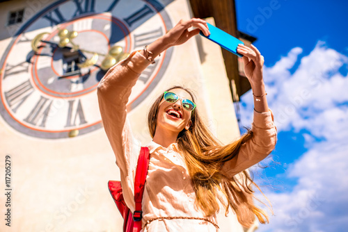 Young female traveler making selfie photo with smart phone on the old clock tower background in Graz town. Traveling in Austria