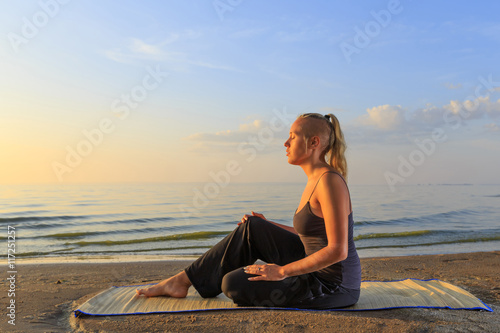 Beautiful young woman practising yoga on mat outdoors at river bank on sand at sunset © yarlander