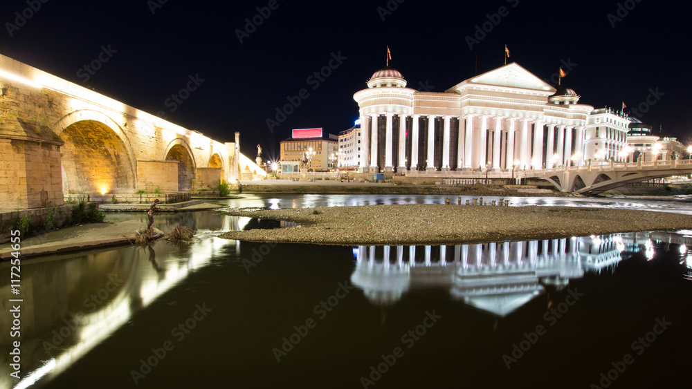 Fototapeta premium Reflections of the Archaeological Museum and the old stone bridge in the river Vardar during the night in Skopje Macedonia