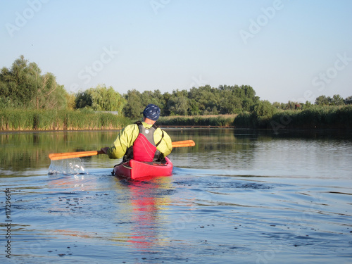 Man paddles a red kayak in summer on river with wooden Greenland paddle