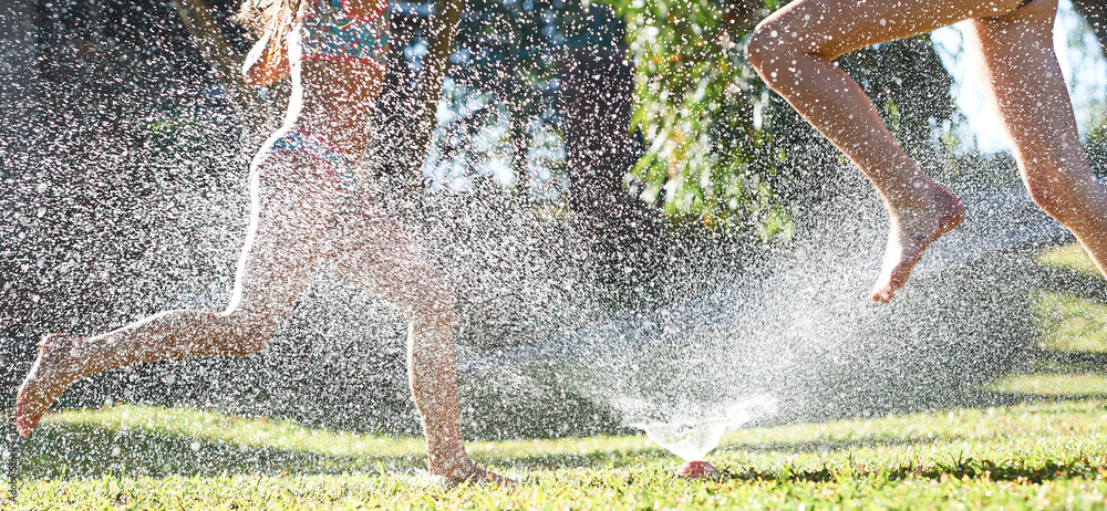 Young girls playing jumping in a garden water lawn sprinkler - obrazy, fototapety, plakaty 