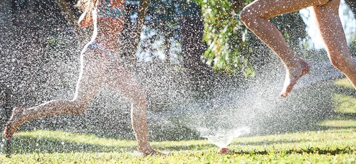 Young girls playing jumping in a garden water lawn sprinkler