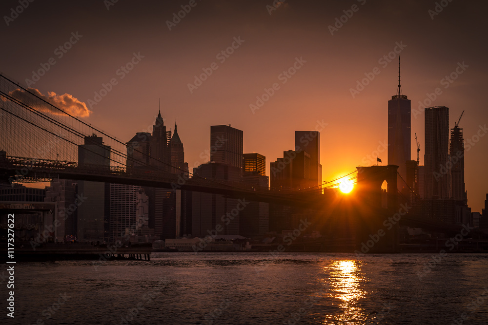 Panorama of a silhouette of Brooklyn Bridge and Manhattan skyline on a clear evening with the sun visible from a narrow passage in between skyscrapers