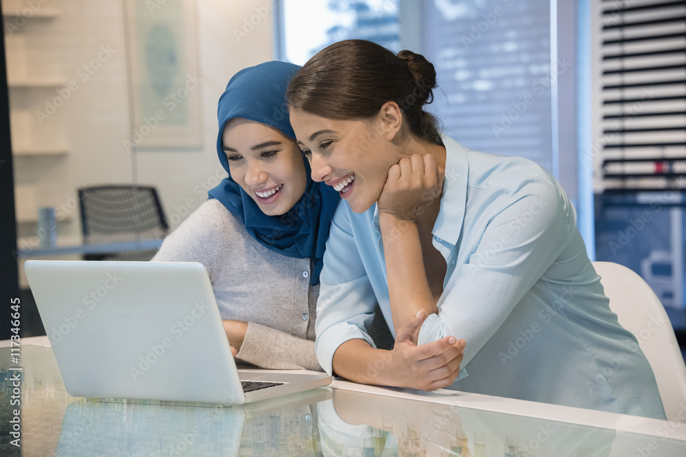 Two happy businesswomen looking at a laptop in an office Two happy businesswomen looking at a laptop in an office
