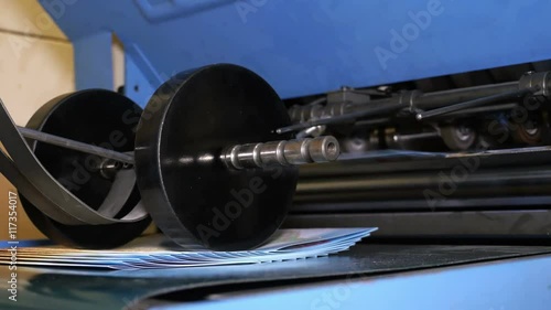 Folded printed sheets of paper fall under working roller, worker collects brochures. Operating folding machine. Related to typography, printing press, printed media. Close up shot. 4K Ultra HD.
