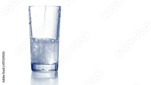 Filling up empty glass with carbonated water. Bubbles go up, drop of water rolls down the side of glass. White background, reflecting surface. Closeup shot. 4K Ultra HD.
