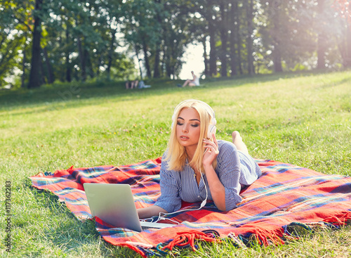 Girl downloading music in her laptop computer at the park. Blonde woman studying outdoors lying on the grass. Beautiful girl in blue shirt lying on a red blanket. © belyjmishka
