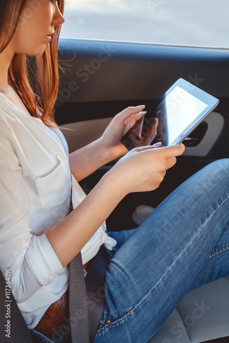 Young, beautiful woman sitting in the back seat of a car with a tablet in hands