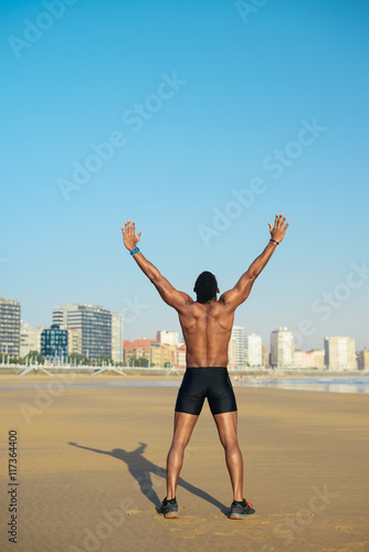 Sport motivation and freedom concept. Back view of strong fit black man raising arms towards the city at the beach for celebrating running workout success.