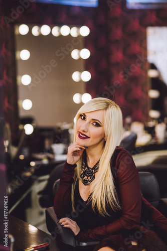 Beautiful fashion blond woman posing near the mirror and gets makeup. Young beautiful model smiling near the mirror. Blonde woman with red lips embracing