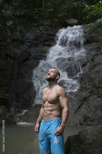 Handsome man with beard wearing blue shorts standing and looking up near waterfall. Male tourist enjoying by a water fall in forest