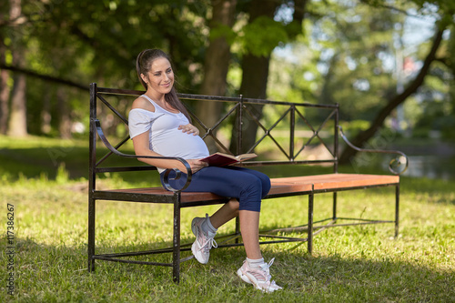 Young pregnant woman with book sits in the park..