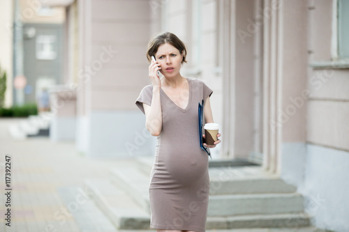 Portrait of unhappy young pregnant business woman walking in the city with document folder and making call. Irritated office worker pregnant woman using smartphone on the street