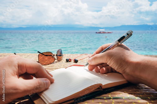 Fototapeta Naklejka Na Ścianę i Meble -  caucasian man is writing sime idea, message or letter in his notepad by pen while he sitting on the beach of tropical sea with boat