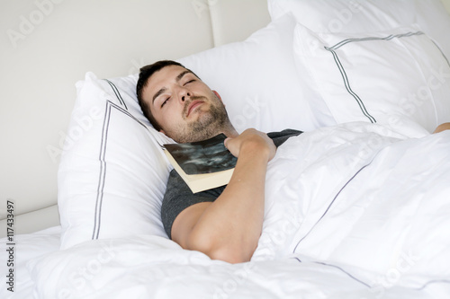 Handsome young man happily sleeping in white bed with book in the hand