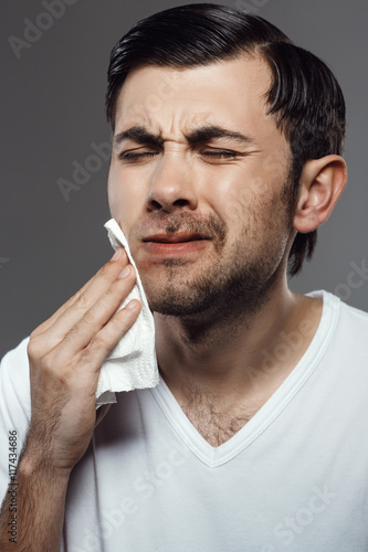 Displeased young man touching face after shaving over grey background.