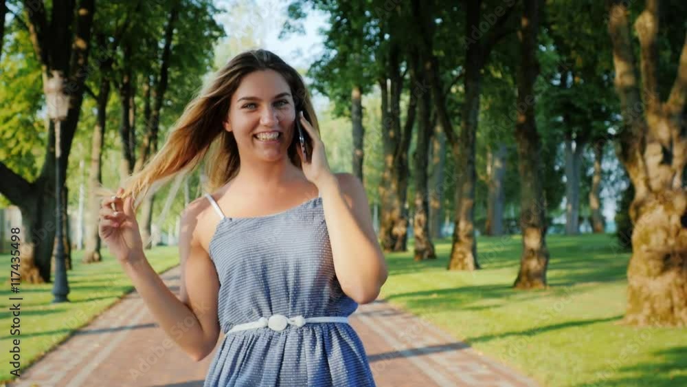 Carefree young woman walking along a path in the park, talking on the phone, flirting: smiling Carefree young woman walking along a path in the park, talking on the phone, flirting: smiling