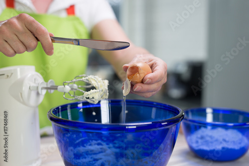 In einer modernen Küche bereitet eine Frau einen Teig zu. Sie schlägt ein Ei auf und gibt es in eine Schüssel. Ein Mixer und eine Schüssel Mehl stehen daneben