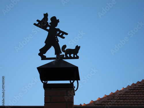 Silhouette of a chimney sweep with cats and stairs to the flue pipe of red brick in front of the blue sky
