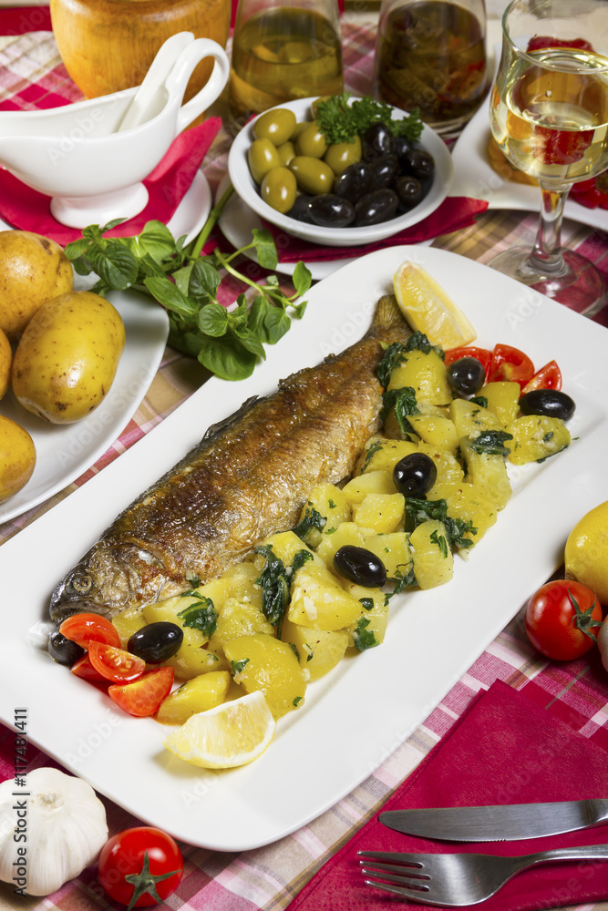 Whole fried trout served on a white plate with potato salad and chard with olives, lemon and cherry tomatoes next to fresh vegetables. Whole fried trout served on a white plate with potato salad and chard with olives, lemon and cherry tomatoes next to fresh vegetables.