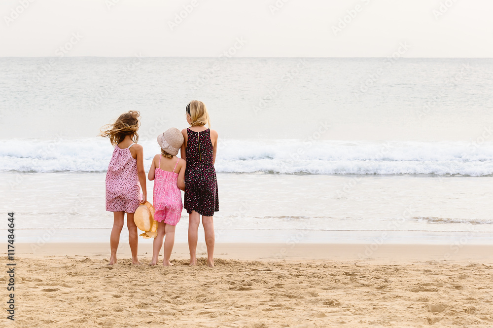 3 sisters stand on beachfront facing the ocean.