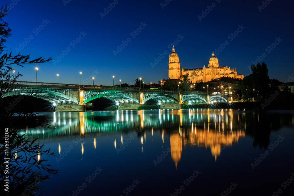 Fototapeta premium Night view of the Cathedral of Salamanca with Enrique Esteban bridge lit foreground and reflections in the Tormes river, Salamanca, Spain