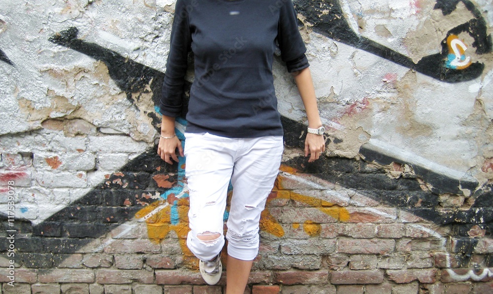 Lifestyle portrait of stylish young girl in casual clothes: black sweater and white ripped jeans standing against colorful urban brick wall background.