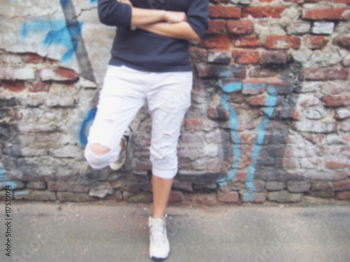 Blurred lifestyle portrait of stylish young man in casual clothes: black sweater, white ripped jeans and sneakers standing against colorful urban brick wall background.