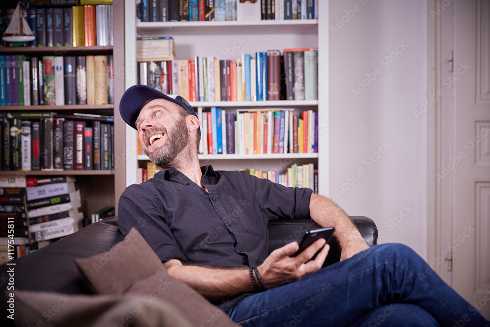 Man sitting in his living room with mobile phone laughing Man sitting in his living room with mobile phone laughing