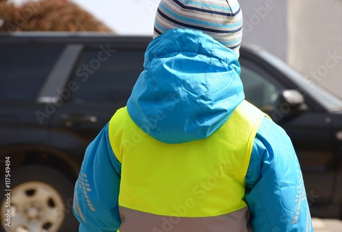 Little boy is going to cross the road where green car is driving. Child is wearing yellow reflective vest and jacket with reflective strips because of safety.