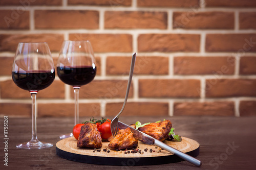 Succulent pieces of grilled pork fillet served with tomatoes branch and lettuce on a board, glass of red wine, knife, fork, cutting the meat on the brick wall background. Organic concept. Horizontal