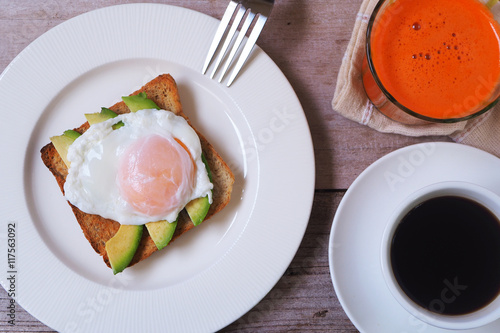 Top view of a breakfast of poached egg and avocado on whole wheat toast with coffee and carrot juice.