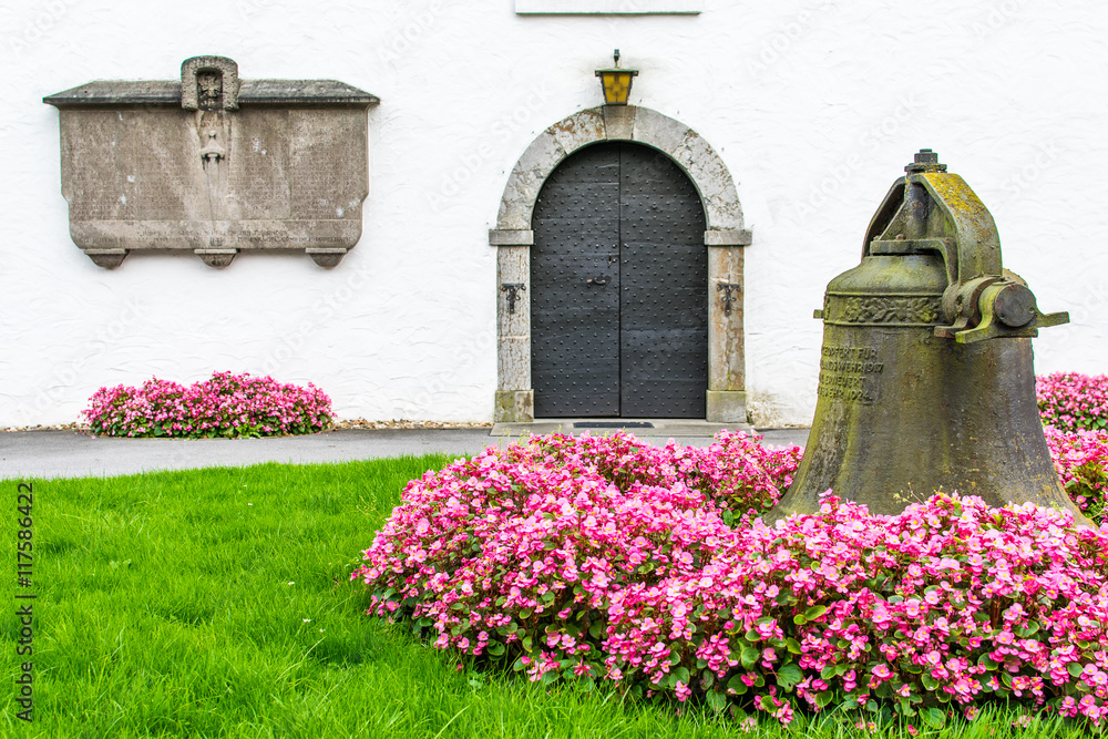 Glocke im Blumenbeet vor kleiner Kapelle