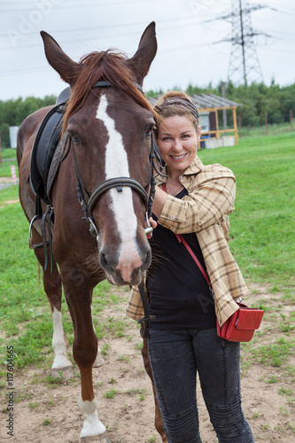 Attractive joyful horsewoman standing near chestnut horse in farm © Kekyalyaynen