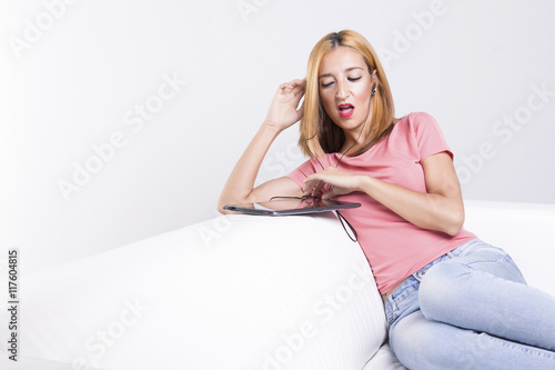 Young woman sitting on a white couch using her tablet pc, feeling disgusted about something on the screen.
