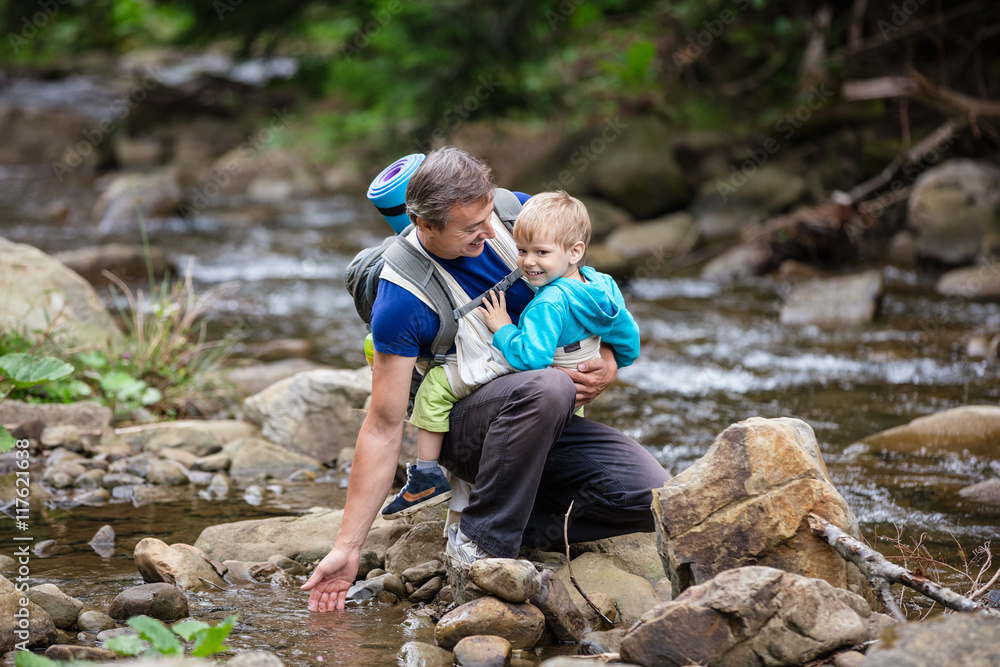 Caucasian man touching water in mountainous river while carrying his son in wrap sling