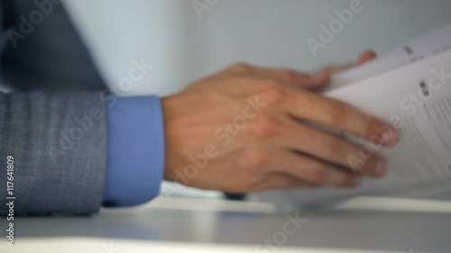 Male notary in suit jacket and light blue shirt holds a document and reads them while sitting at table, closeup.