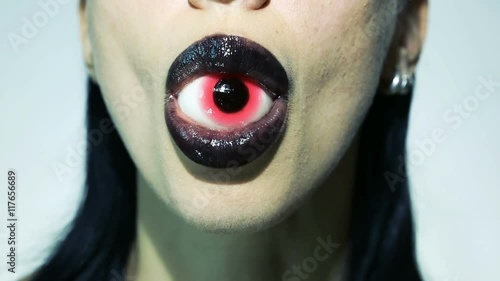 Horror psychedelic concept - Beautiful Sexy Stylish Woman Eating Red Eye and licking her dark lips with tongue. Halloween ClubTrendy look. Emotional Vampire Girl. Lips with black lipstick close up. 