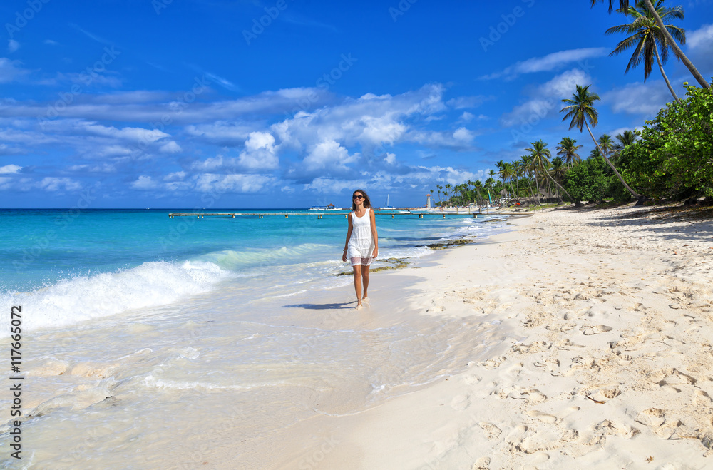 Young woman walking along white sand tropical beach. Beautiful girl in white dress on the beach. Travel and vacation. Freedom concept.