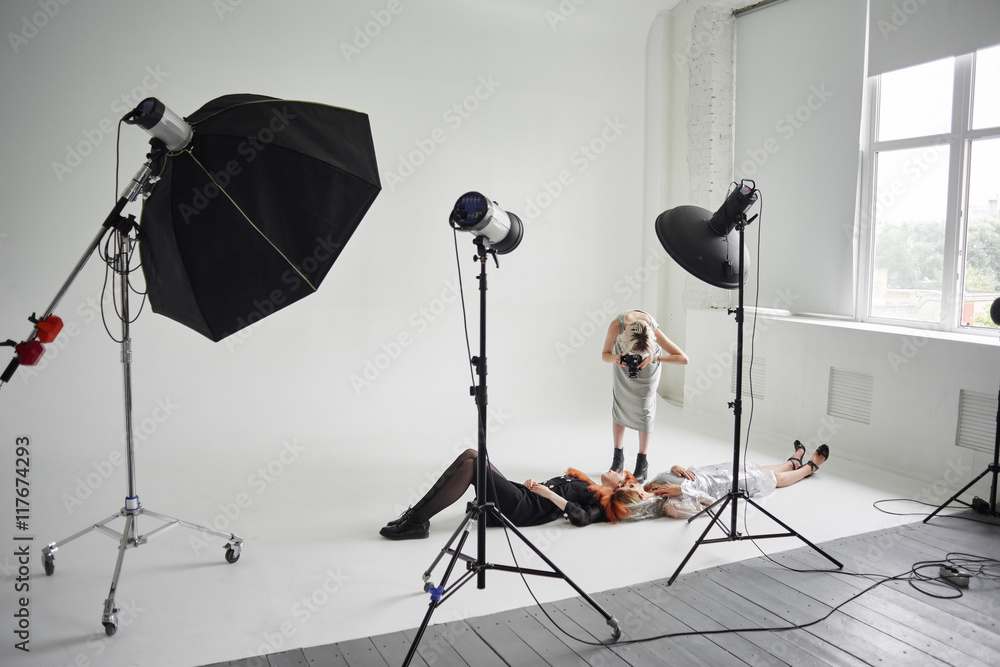 Girl photographer photographing fashion models in a clear coat with a pomegranate mask on the face and black clothes lying on white floor in Studio