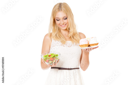Half-length portrait of very beautiful woman holding cake and fresh vegetables. Young housewife choosing sweets or healthy eating - cake and salad. Laughing at camera. Isolated on white background.