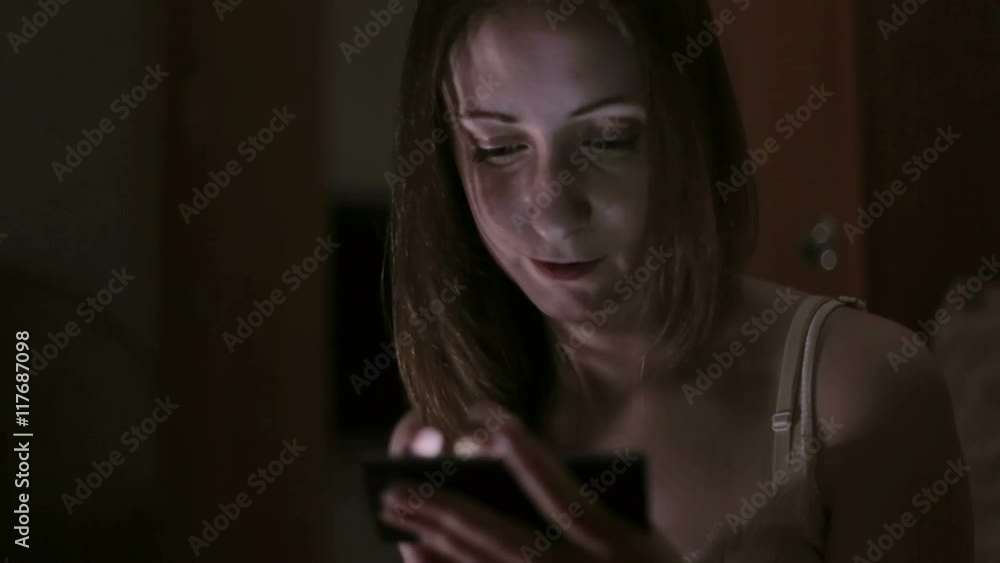 Attractive, young woman browsing photos or internet on smartphone lying in bed at night Attractive, young woman browsing photos or internet on smartphone lying in bed at night