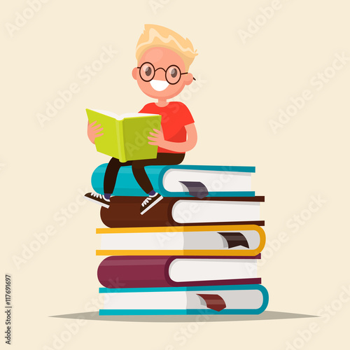 Boy with glasses reading a book sitting on a stack of textbooks.
