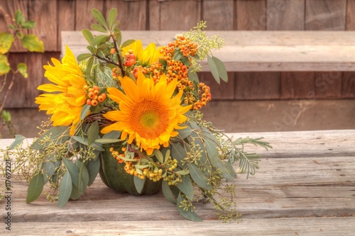 Fototapeta Naklejka Na Ścianę i Meble -  Holiday flower bouquet inside a gourd vase with red berries, green leaves and sprigs, and sunflowers on a rustic wood farm house background. 