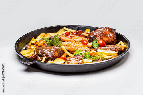 Meat assortment on a large frying pan, potato slices braised cabbage and tomatoes. On a white background